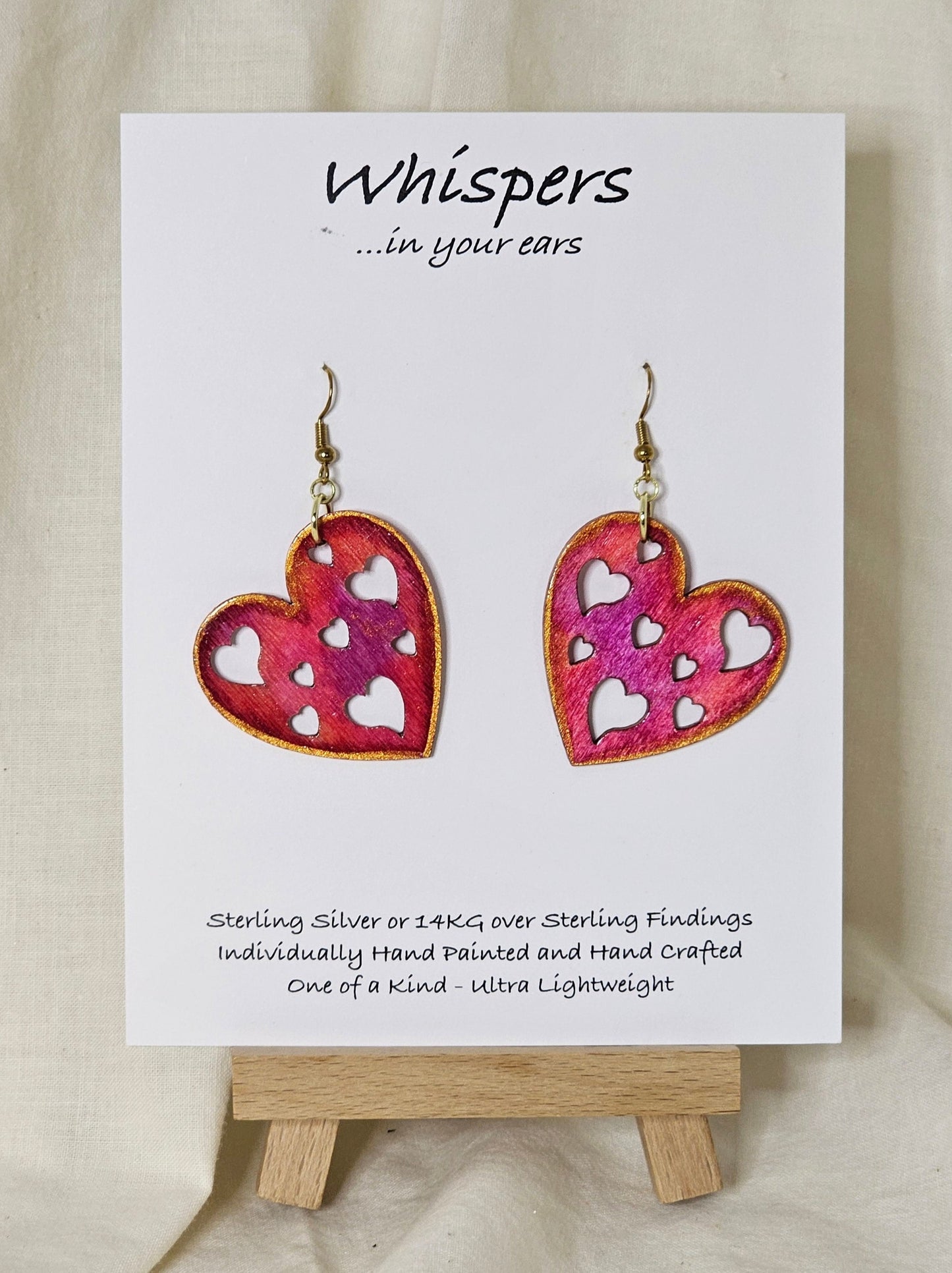 Heart Earrings Wood die cut hand painted Ultra lightweight. Heart shape cut outs. Painted variations of pinks. Gold edge detail. Enameled for durability. Back is painted in the same color and pattern. Heart is 1 1/2 " in width. 14 kg over silver findings. Hangs 1 1/2" in length