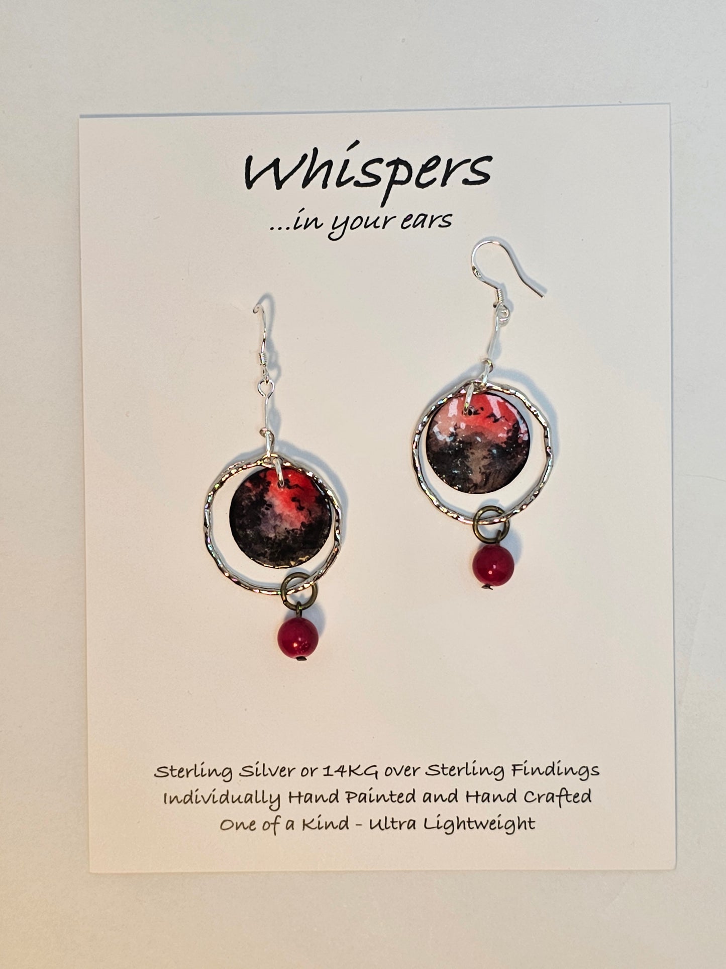 Red Skies Hand Painted Paper and Silver Alloy Earrings. 3/4" Circular watercolor painting of trees and mountains in silhouette against a red sky. Painting hanging in the center of a silver alloy circle. below hangs a red bead embellishment. Back is painted to match. Sterling Silver findings. Resin coating for durability. Hangs 2 1/8" in length