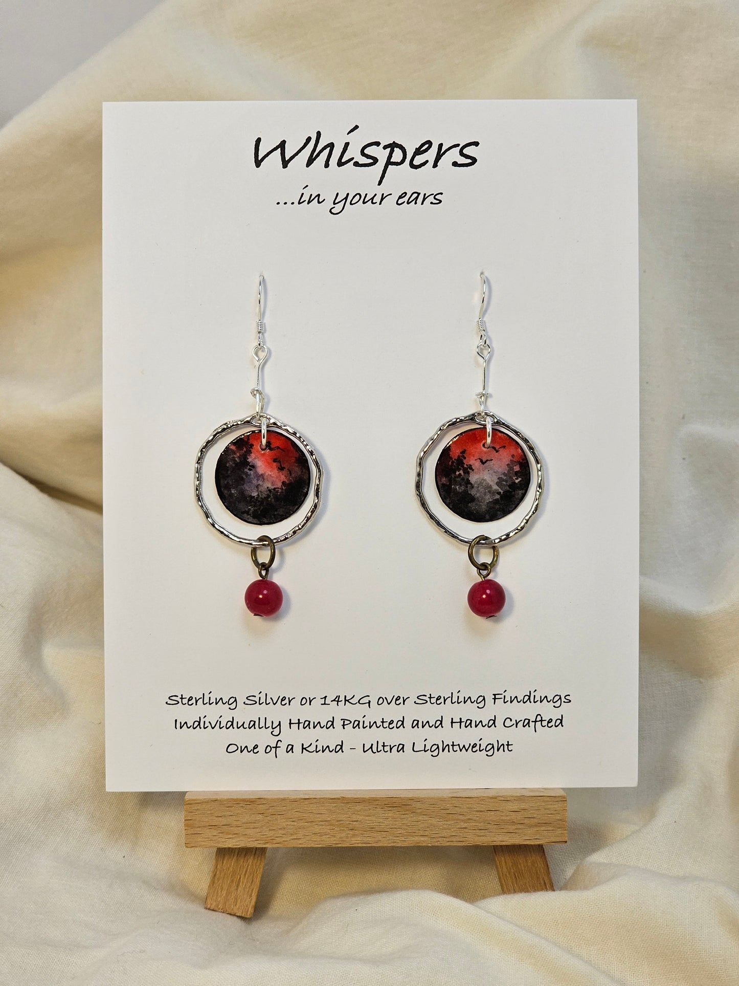 Red Skies Hand Painted Paper and Silver Alloy Earrings. 3/4" Circular watercolor painting of trees and mountains in silhouette against a red sky. Painting hanging in the center of a silver alloy circle. below hangs a red bead embellishment. Back is painted to match. Sterling Silver findings. Resin coating for durability. Hangs 2 1/8" in length