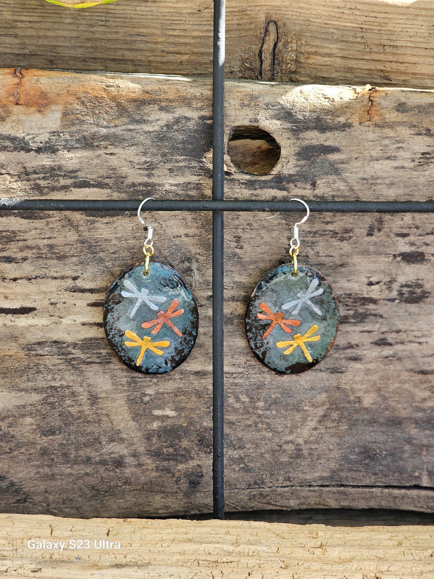 Hand painted Watercolor Mixed Media Collage Dragonfly Trinity. Ultra lightweight paper earrings. Grey blue patina background.  Layered collage with copper, silver and gold painted paper dragonflies.  Edged in dark brown patina. Back is painted in complimentary color. Oval in shape. Combination of14kg over sterling silver  and Sterling silver findings. Hangs 1 3/4" in Length