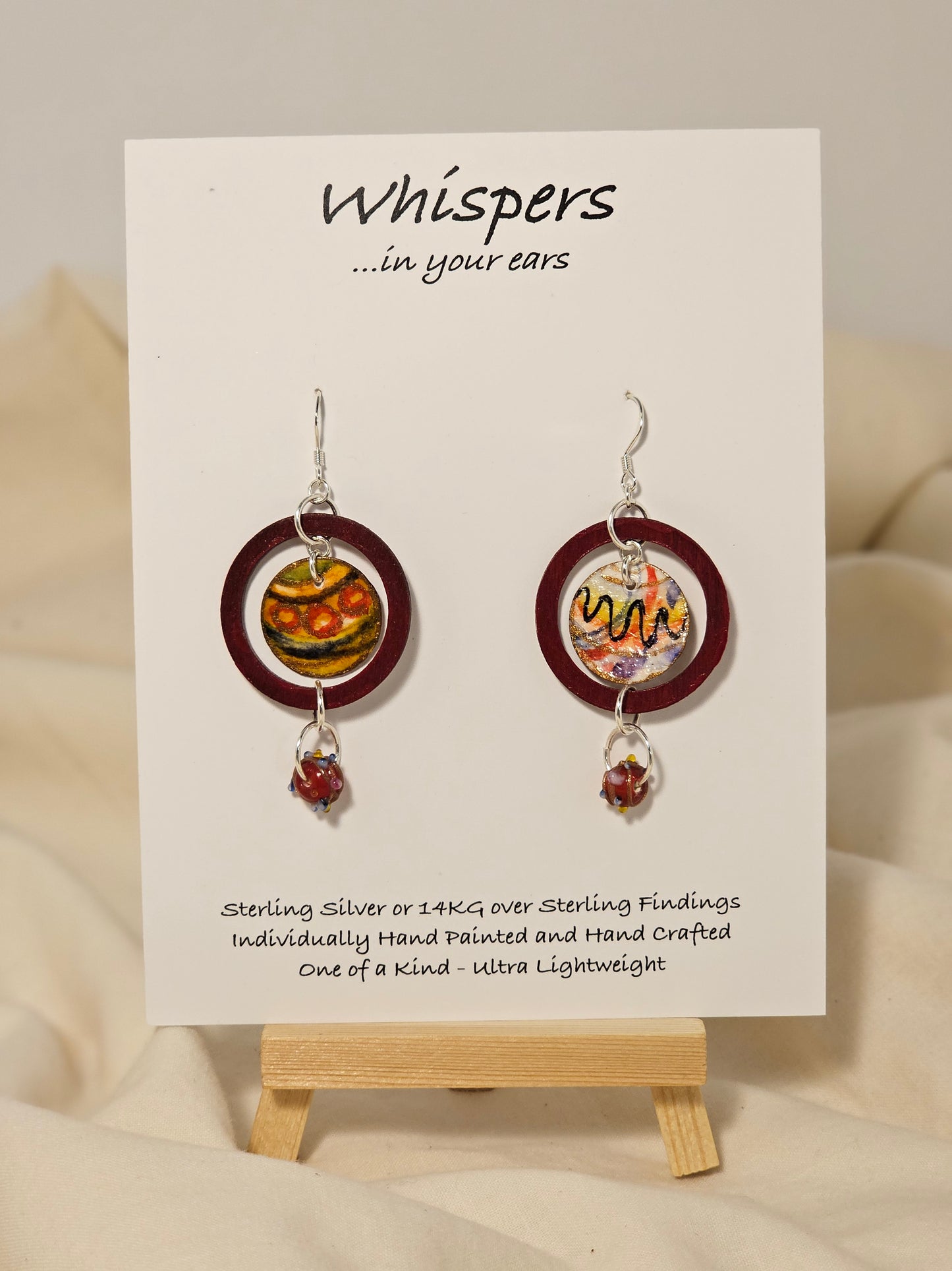 Red Ornament Ring Wood and Paper Earrings. 1 1/4" painted red wood ring with hanging hand painted watercolor ornament in the center. Multicolored glass bead hanging at the bottom. Back is painted in complimentary design. Enameled for durability. Sterling Silver findings. Hangs 2" in length
