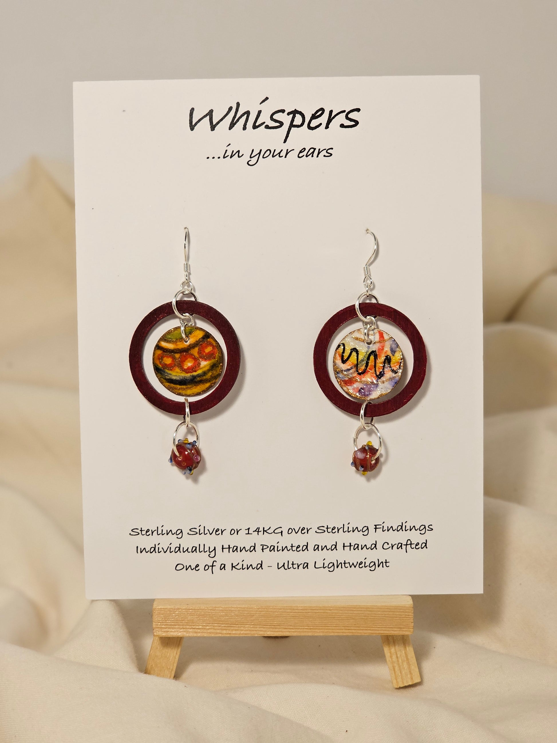 Red Ornament Ring Wood and Paper Earrings. 1 1/4" painted red wood ring with hanging hand painted watercolor ornament in the center. Multicolored glass bead hanging at the bottom. Back is painted in complimentary design. Enameled for durability. Sterling Silver findings. Hangs 2" in length