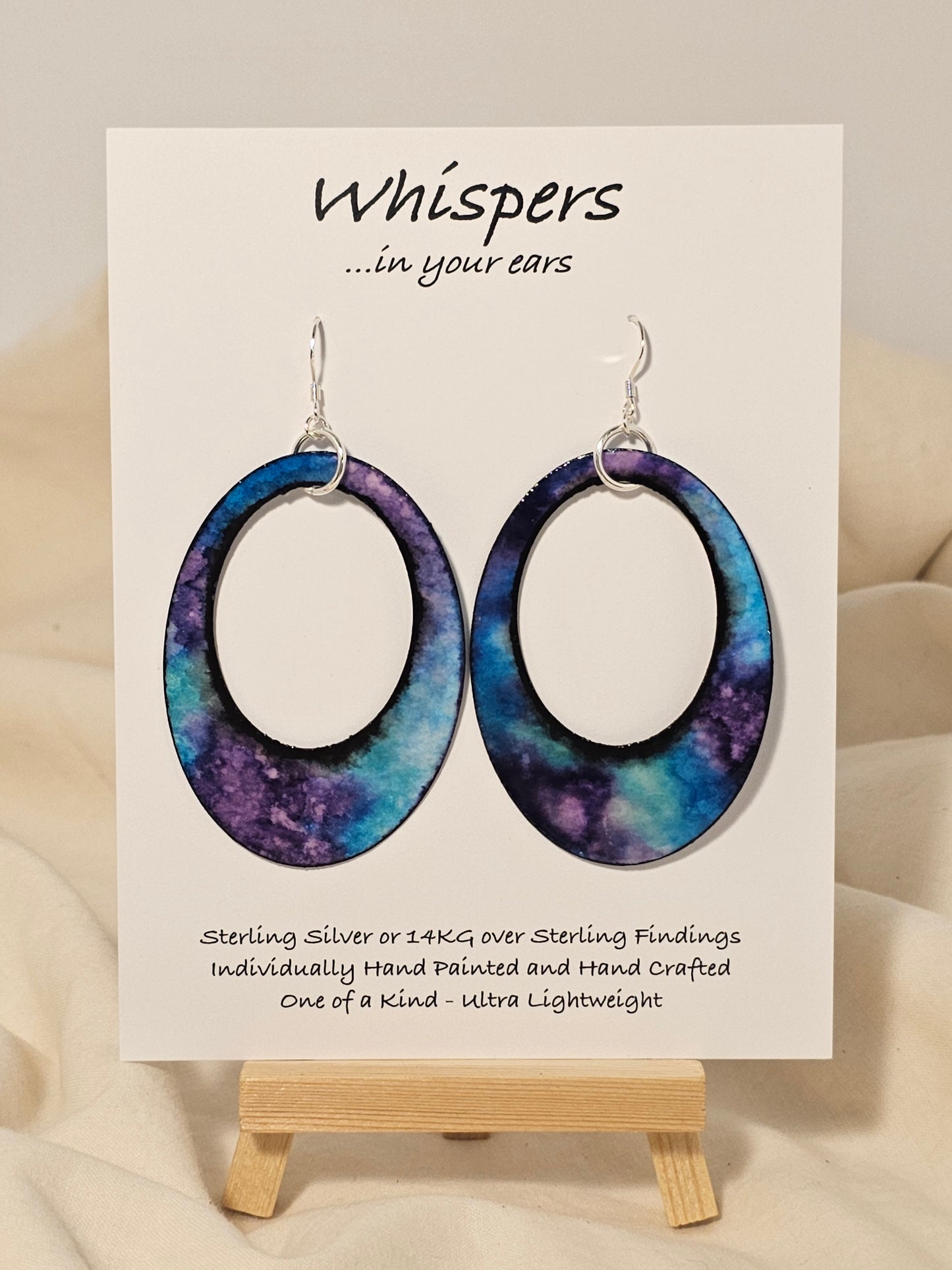 Galaxy Statement Hand Painted Paper Earrings. 2 1/2" Oval with cut out.&nbsp; Statement. Watercolor painted turquoise blue, aqua and purple. Back is painted to match. Edged in black. Resin coating for durability. Sterling Silver findings. Hangs 2 3/4" in length