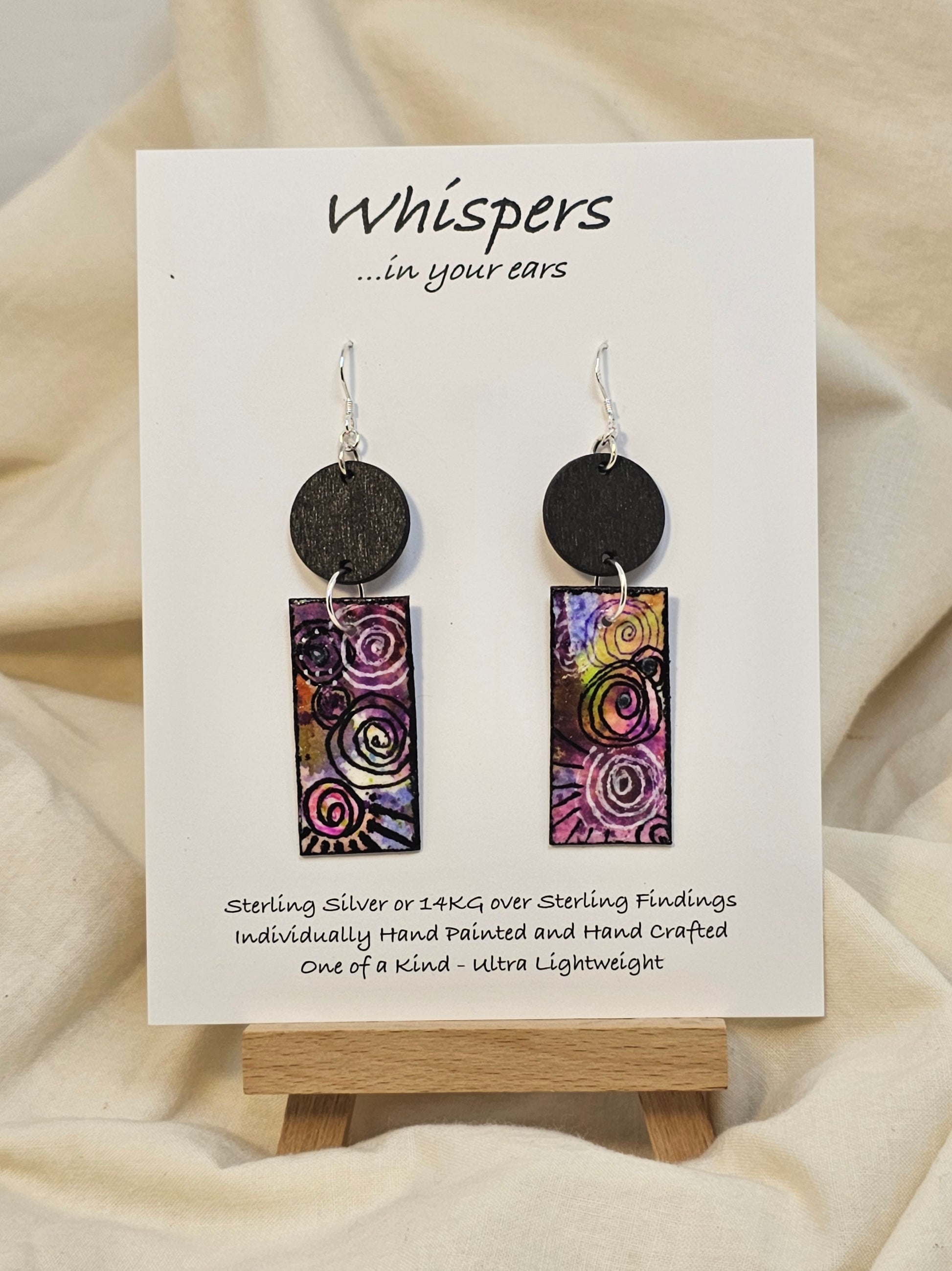 Black Spiral Motif Hand Painted Paper and Wood Earrings. 3/4" x 1 5/8" Paper Rectangle hand painted brightly colored background with black swirl accents. Edged in black. Hanging from embellished wooden disc painted black. Back is painted to match. Resin coating for durability. Sterling Silver findings. Hangs 2 1/2" in length