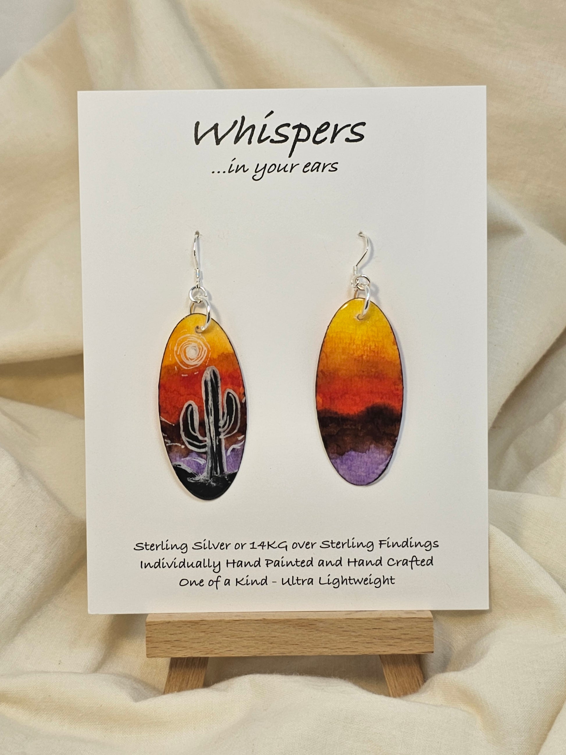Desert Sunset Hand Painted Paper Earrings.&nbsp; Ultra lightweight 2" Elongated oval. Features Cactus in silhouette. Vibrant orange yellow and purple sky. Painted dark edge. Back is painted with similar color. Resin coating for durability. Sterling Silver findings. Hangs 2 1/8" in length
