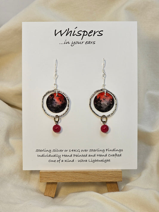 Red Skies Hand Painted Paper and Silver Alloy Earrings. 3/4" Circular watercolor painting of trees and mountains in silhouette against a red sky. Painting hanging in the center of a silver alloy circle. below hangs a red bead embellishment. Back is painted to match. Sterling Silver findings. Resin coating for durability. Hangs 2 1/8" in length