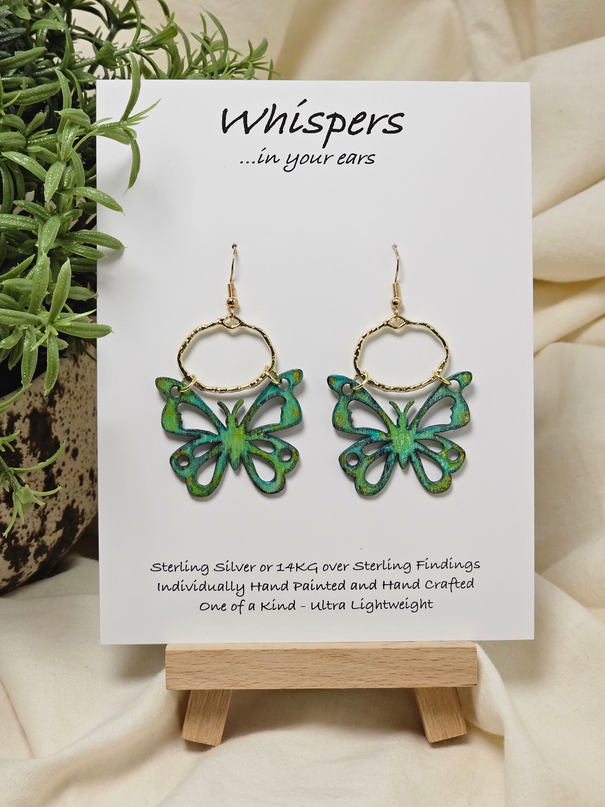 Aqua and Lime Butterfly Hand Painted Wood Earrings. 1"x 1 1/2" Die cut butterfly painted in greens and blues with gold edge accents. Back of butterfly painted in similar design. Hangs gently from gold alloy oval. Hangs 1 5/8". Enameled for durability. 14kg over sterling silver ear wires and findings. Ultra light weight