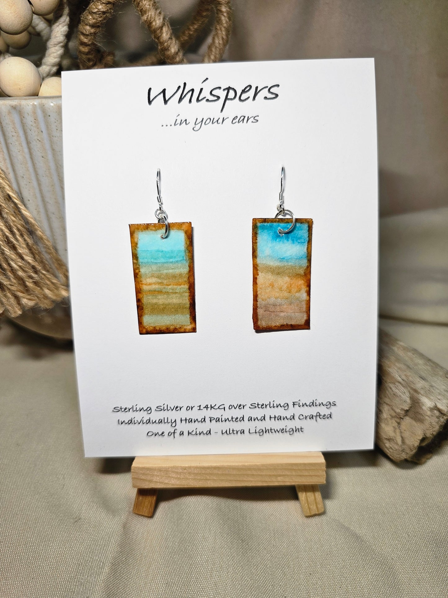 Beach Impressions Hand Painted Paper Earrings. Ultra lightweight 3/4" x 1 1/2" rectangle painted with watercolor. Depicts impressionistic beachscape. Subtle ocean color accents. Edges painted in brown to frame the scene. Back is painted with the same design. Resin coating for durability. Sterling silver ear wires. Hangs 1 3/4" in length