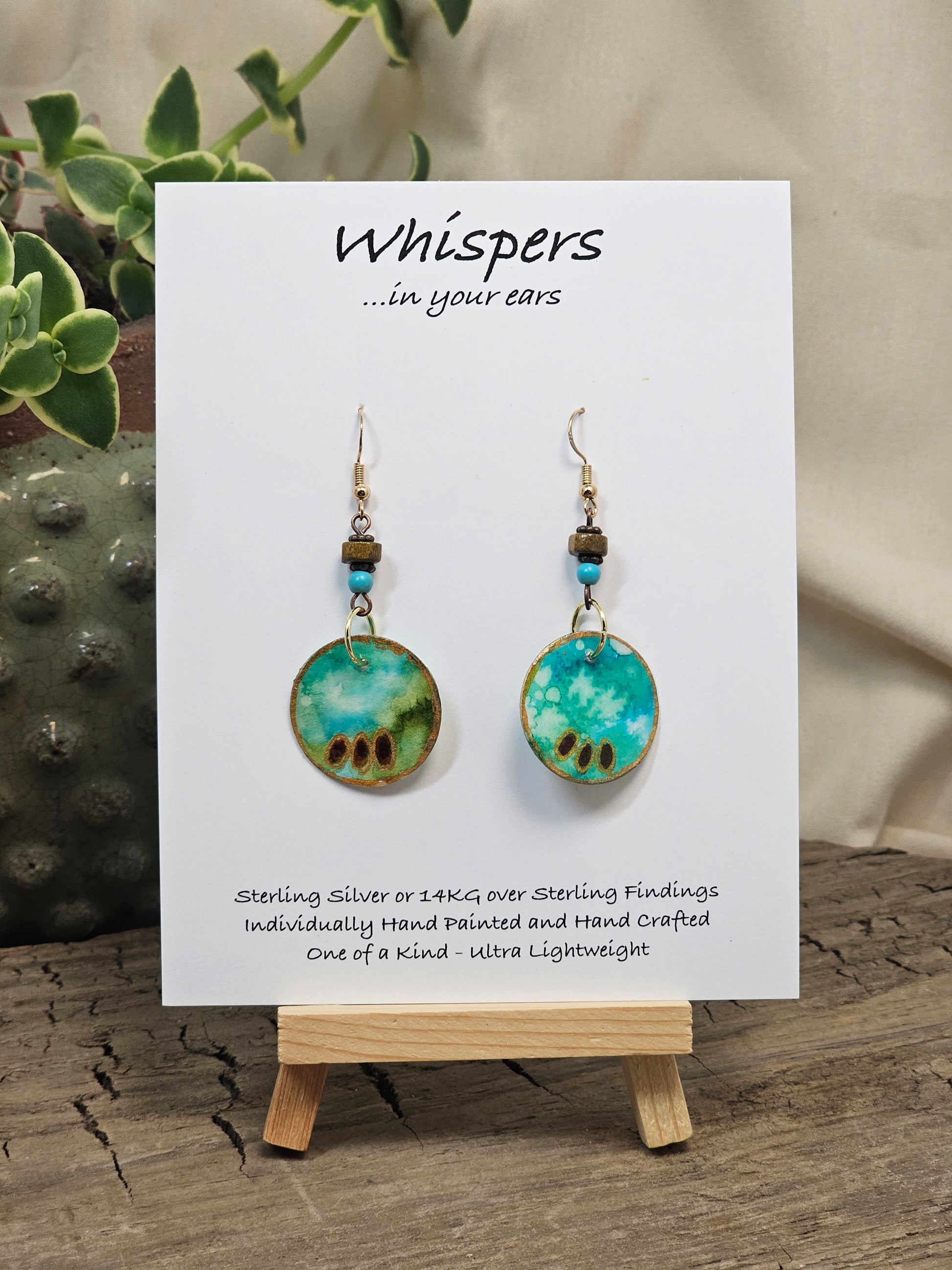 Aqua Egyptianesque Hand Painted Paper Earrings. 1" Circular artisan paper aqua blue, moss green and brown oval pattern with metallic painted edges. Hangs from metal decorative wooden and turquoise blue beads, Resin Coating for durability. Back is done to compliment. 14kg over Sterling silver ear wires and sterling silver findings. Hangs 1 3/4"