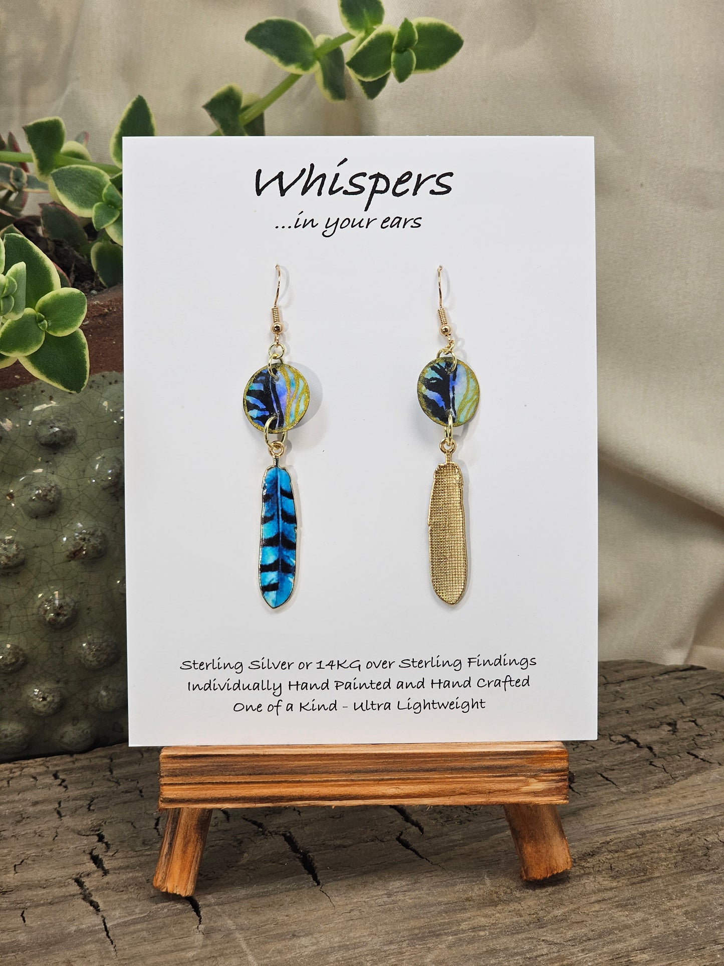 Black Feathers Hand Painted Paper Earrings. 5/8" circular hand painted paper earring. Below hangs a gorgeous light weight metal enameled feather charm pendant. Back is gold in color. Aqua blue, royal blue, black and white, metallic accents edged in gold. Back of paper is painted in similar design and colors. Resin coated for durability. 14kg over sterling silver ear wires and findings. Hangs 2 1/2" in length