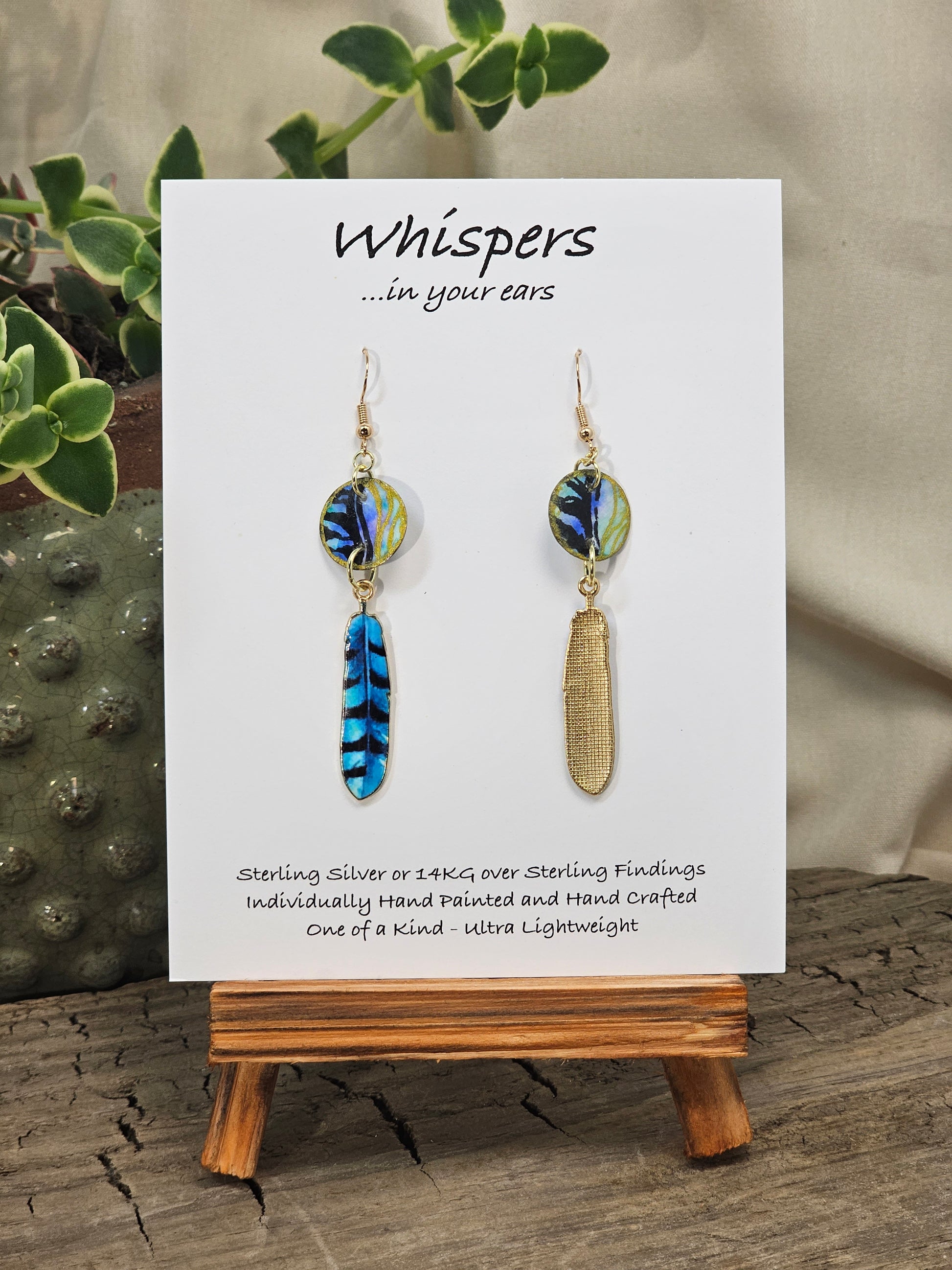 Black Feathers Hand Painted Paper Earrings. 5/8" circular hand painted paper earring. Below hangs a gorgeous light weight metal enameled feather charm pendant. Back is gold in color. Aqua blue, royal blue, black and white, metallic accents edged in gold. Back of paper is painted in similar design and colors. Resin coated for durability. 14kg over sterling silver ear wires and findings. Hangs 2 1/2" in length