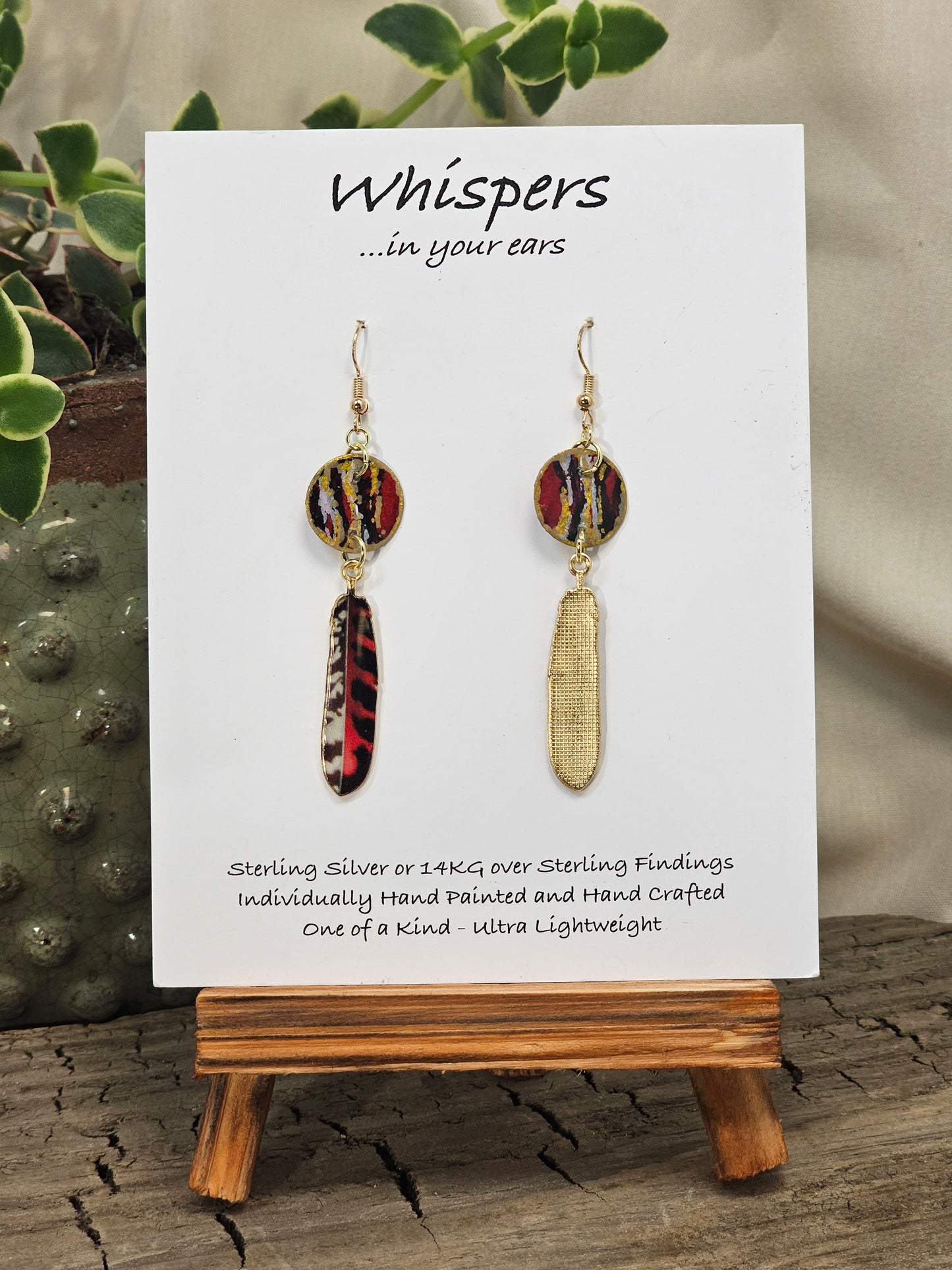 Tiger Striped Feather Hand Painted Paper Earrings. 5/8" circular hand painted paper earring. Below hangs a gorgeous light weight metal enameled feather charm pendant. Back is gold in color. Red brown, gold, black and white, metallic accents edged in gold. Back of paper is painted in similar design and colors. Resin coated for durability. 14kg over sterling silver ear wires and findings. Hangs 2 1/2" in length