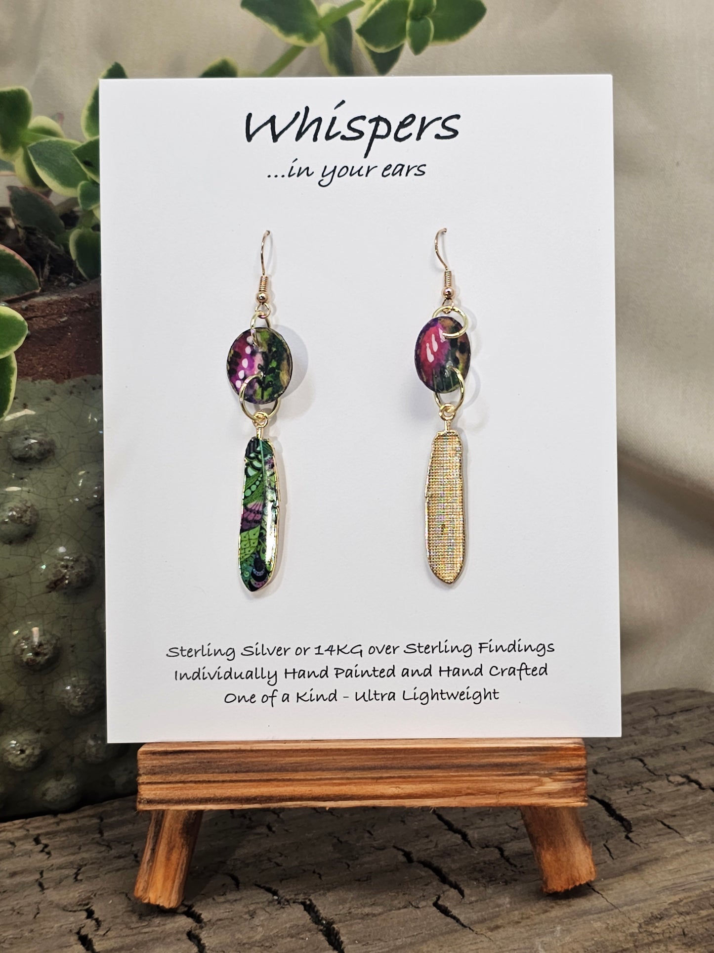 Jungle Pattern Feather Hand Painted Paper Earrings. 5/8" circular hand painted paper earring. Below hangs a gorgeous light weight metal enameled feather charm pendant. Back is gold in color. Pink, purple, gold, green, black and white, metallic accents edged in gold. Back of paper is painted in similar design and colors. Resin coated for durability. 14kg over sterling silver ear wires and findings. Hangs 2 1/2" in length