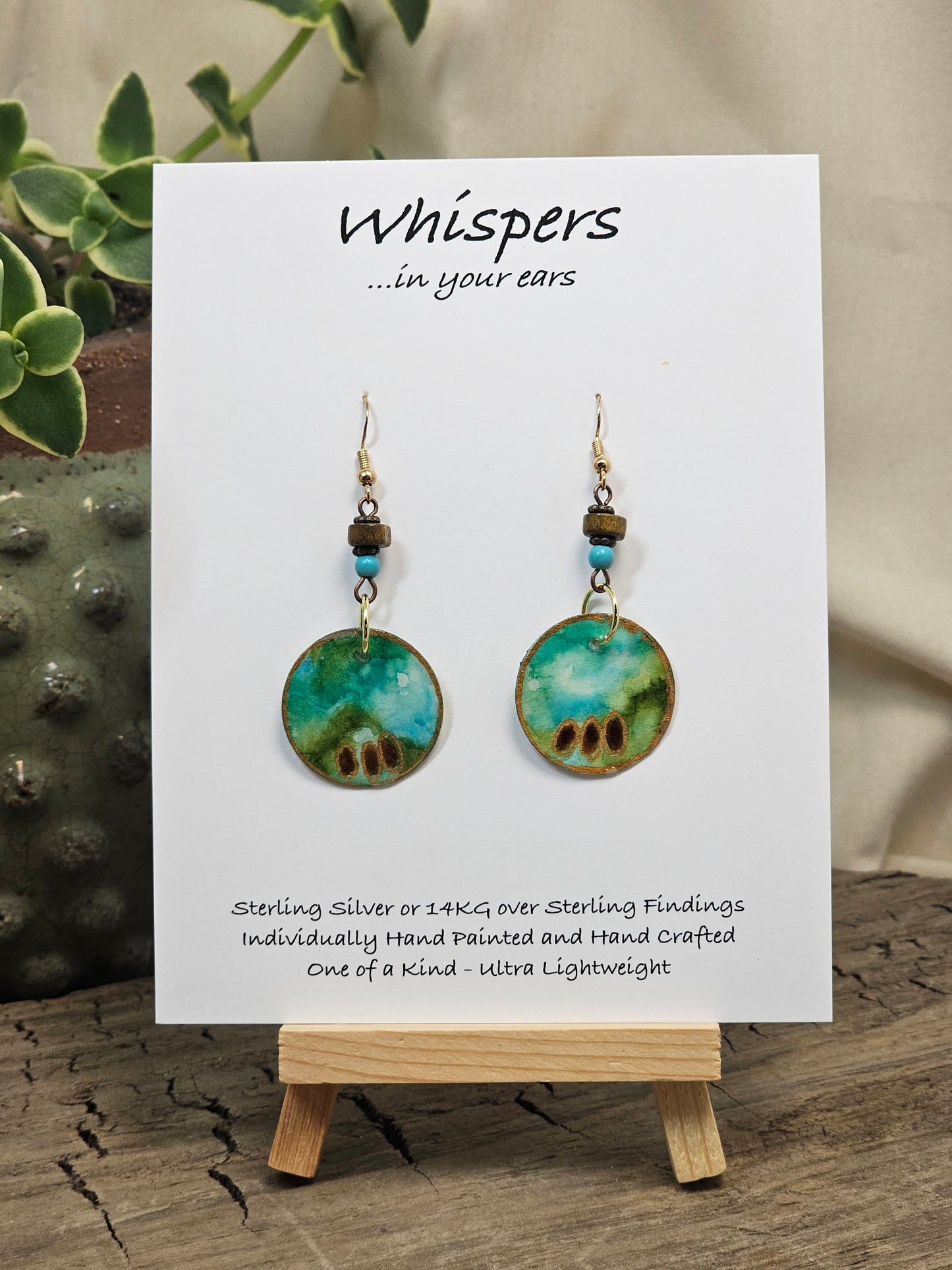Aqua Egyptianesque Hand Painted Paper Earrings. 1" Circular artisan paper aqua blue, moss green and brown oval pattern with metallic painted edges. Hangs from metal decorative wooden and turquoise blue beads, Resin Coating for durability. Back is done to compliment. 14kg over Sterling silver ear wires and sterling silver findings. Hangs 1 3/4"