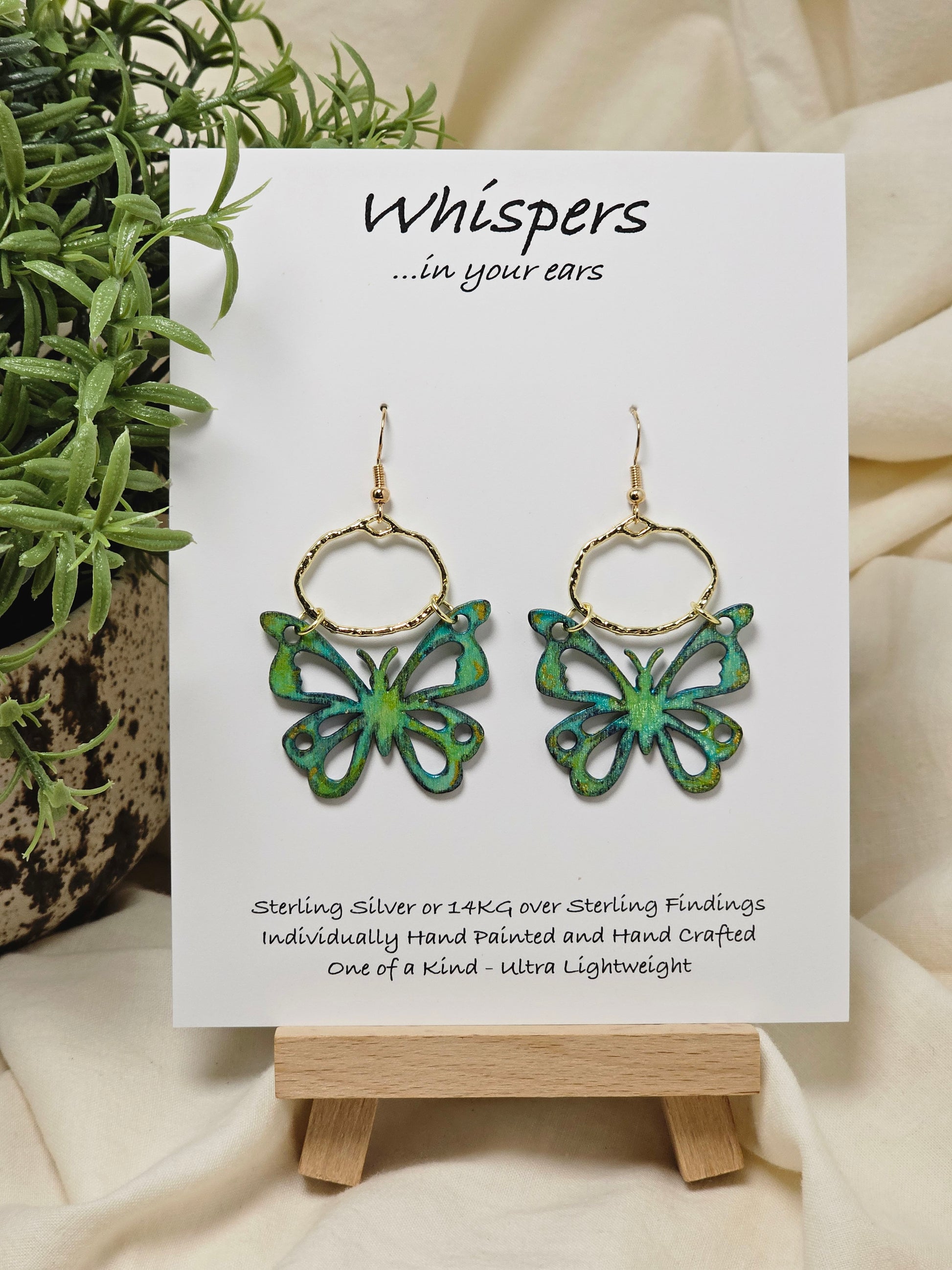 Aqua and Lime Butterfly Hand Painted Wood Earrings. 1"x 1 1/2" Die cut butterfly painted in greens and blues with gold edge accents. Back of butterfly painted in similar design. Hangs gently from gold alloy oval. Hangs 1 5/8". Enameled for durability. 14kg over sterling silver ear wires and findings. Ultra light weight