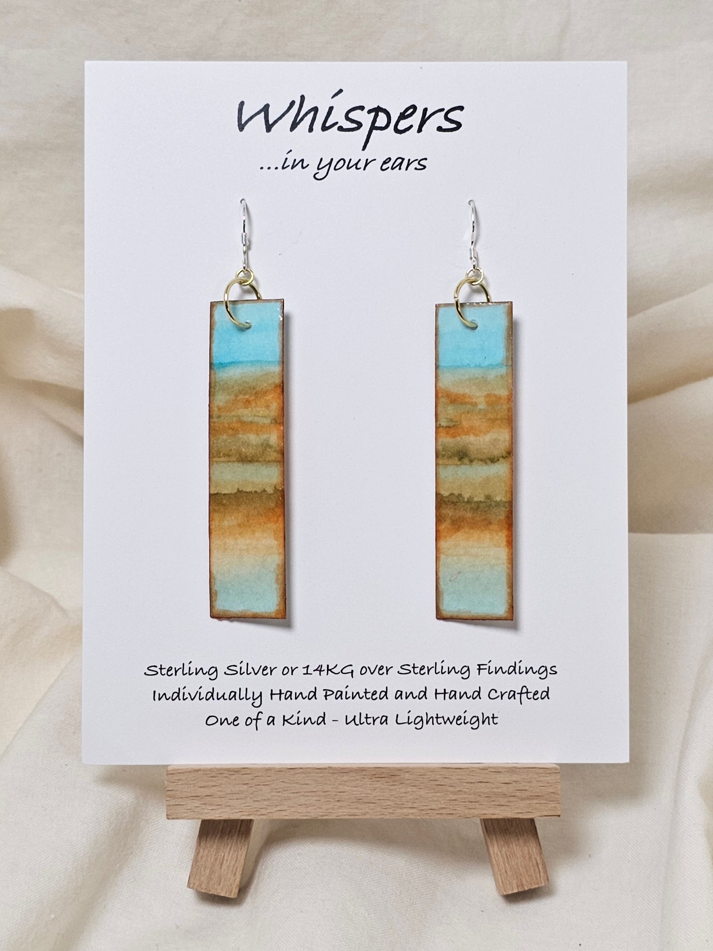 Beachscape Reflection Statement Hand Painted Paper Earrings. Abstract beachscape painted in and on heavy watercolor paper. 1/2" x 2 1/2" elongated rectangle. In muted blues and beiges and corals. Reflection of landscape. Resin coating for durability. Sterling silver ear wires and findings. Ultra lightweight. Hangs 2 3/4"