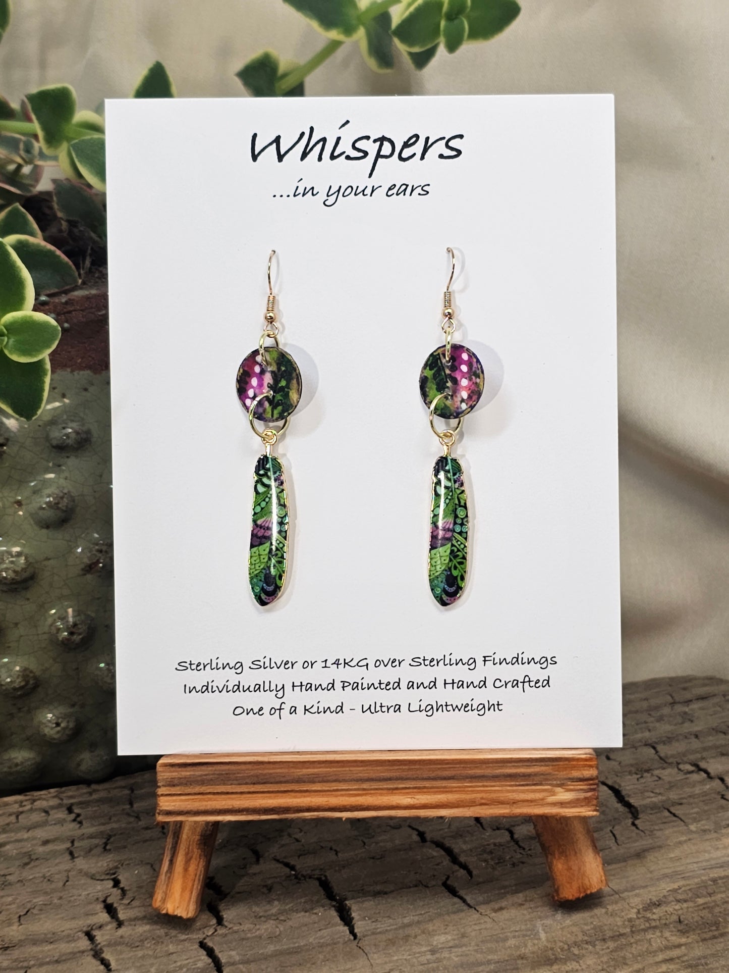 Jungle Pattern Feather Hand Painted Paper Earrings. 5/8" circular hand painted paper earring. Below hangs a gorgeous light weight metal enameled feather charm pendant. Back is gold in color. Pink, purple, gold, green, black and white, metallic accents edged in gold. Back of paper is painted in similar design and colors. Resin coated for durability. 14kg over sterling silver ear wires and findings. Hangs 2 1/2" in length