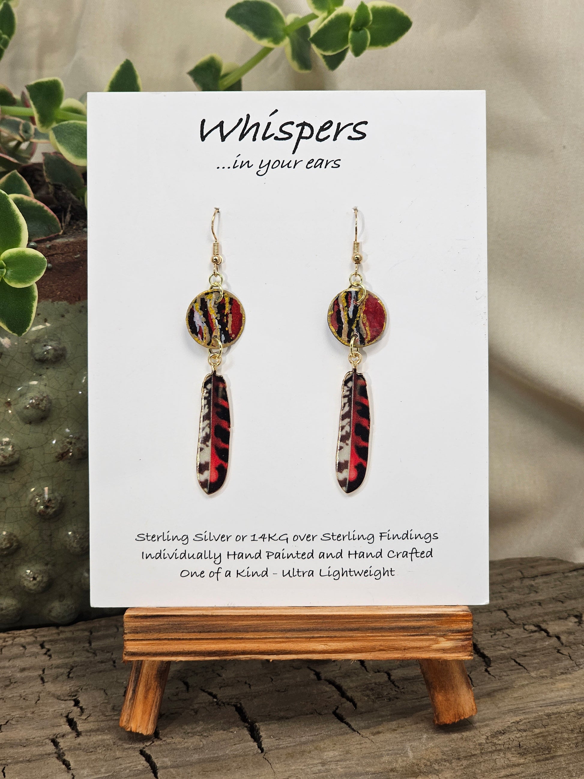 Tiger Striped Feather Hand Painted Paper Earrings. 5/8" circular hand painted paper earring. Below hangs a gorgeous light weight metal enameled feather charm pendant. Back is gold in color. Red brown, gold, black and white, metallic accents edged in gold. Back of paper is painted in similar design and colors. Resin coated for durability. 14kg over sterling silver ear wires and findings. Hangs 2 1/2" in length